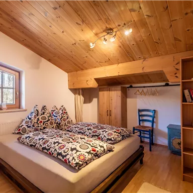 A cozy bedroom with a wooden roof and a large bed. A writing chair and a shelf complement the space.