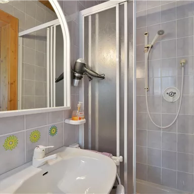 Ein Badezimmer mit Dusche, Waschbecken und Spiegel. Die Wände sind hell gefliest, und es gibt farbenfrohe Dekorationen.