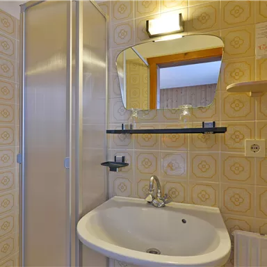 Ein kleines Badezimmer mit einer Dusche, einem rechteckigen Waschbecken und einem Spiegel. Die Wände sind mit gelben Fliesen dekoriert.