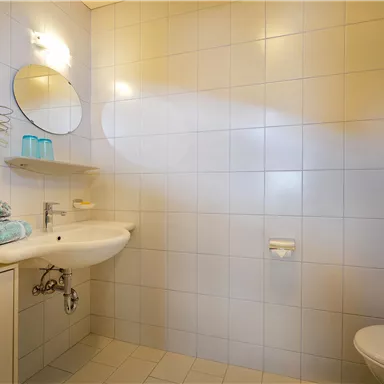 A modern bathroom with white tiles and a shower. Fresh towels are laid on the sink and there is a large mirror.