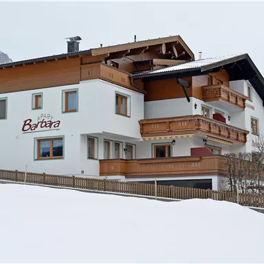 Ein gemütliches Haus im alpinen Stil, umgeben von Schnee. Im Hintergrund sind Berge sichtbar, was eine malerische Winterlandschaft schafft.