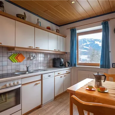 A modern kitchen with white cabinets and a wooden ceiling. Through the window, there is a view of the mountains.