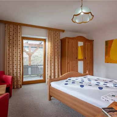 A cozy bedroom with a double bed and a large window. The room is bright and friendly, furnished with a wooden wardrobe and a small table.