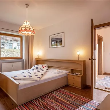 A cozy bedroom with a wooden bed and colorful carpet. Natural light flows through the window, creating an inviting atmosphere.