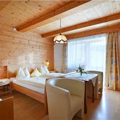 A cozy room with wooden walls, two beds, and a table. Large windows provide plenty of daylight.