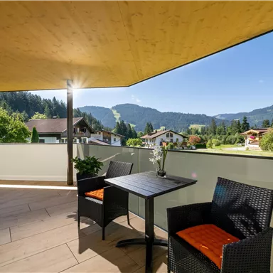 Eine sonnige Terrasse mit einem Tisch und zwei Stühlen. Im Hintergrund sind grüne Hügel und eine klare blaue Himmel zu sehen.