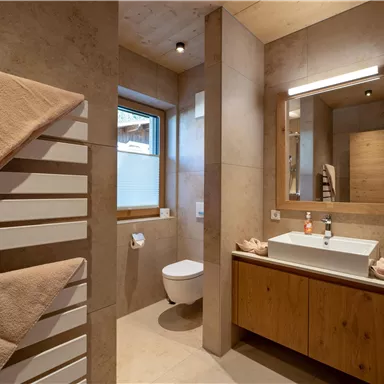 Ein modernes Badezimmer mit Holz- und Steinakzenten. Es verfügt über ein Waschbecken, eine Toilette und große Fenster für viel Licht.