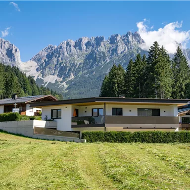 Ein modernes Haus umgeben von Natur, mit Bergen im Hintergrund. Der Himmel ist klar und die Landschaft ist grün und weitläufig.