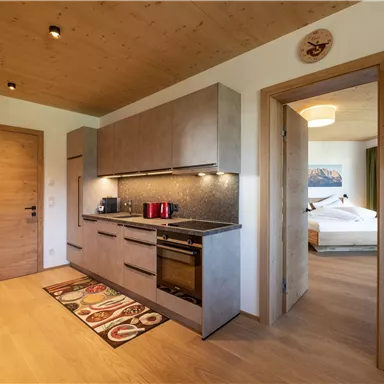 Eine moderne Küche mit grauen Schränken und hochwertigen Geräten. Im Hintergrund ist ein gemütlicher Wohnbereich sichtbar.