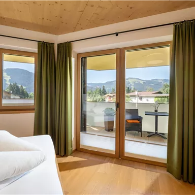 Ein gemütliches Zimmer mit großen Fenstern und Blick auf die Berge. Der Raum ist hell und verfügt über grüne Vorhänge sowie einen Balkon.