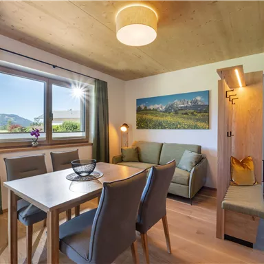 Ein gemütlicher Wohnbereich mit einem Tisch und Stühlen, einem Sofa und viel natürlichem Licht. Der Blick aus dem Fenster zeigt eine schöne Landschaft mit Bergen im Hintergrund.