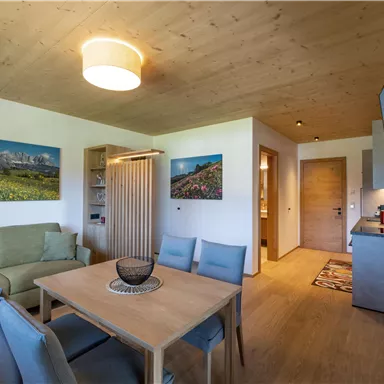 Ein modernes Wohnzimmer mit einem Essbereich und einer gemütlichen Sofaecke. An den Wänden hängen Bilder von Landschaften, und die Küche ist gut ausgestattet.