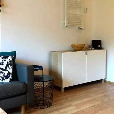 Ein modernes Wohnzimmer mit einem grauen Sofa und einem Beistelltisch. Auf der gegenüberliegenden Seite steht ein weißer Schrank und ein dekorativer Korb.