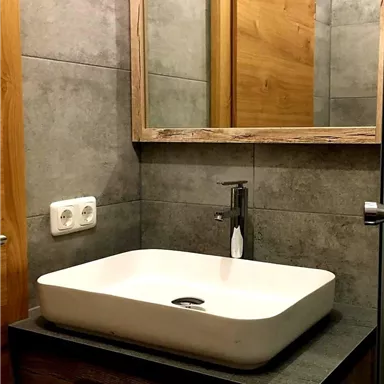 Ein modernes Badezimmer mit einer eleganten Weißkeramikschüssel und einem Holzunterschrank. An der Wand befindet sich ein Spiegel, und die Wände sind mit grauen Fliesen verkleidet.