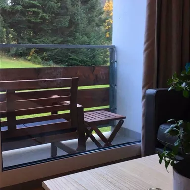Ein gemütlicher Wohnraum mit Blick auf einen Balkon und grüne Bäume. Der Tisch ist aus Holz und eine Pflanze steht darauf.
