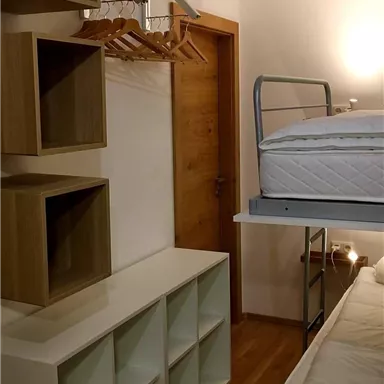 Ein einfaches Zimmer mit Etagenbett, Regalen und einem Kleiderständer. Der Boden ist aus Holz und die Wände sind hell gestrichen.
