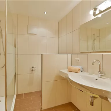 Ein modernes Badezimmer mit einer Dusche und hellen Fliesen. Auf dem Waschbecken liegen ein Handtuch und ein orangefarbenes Handtuch.