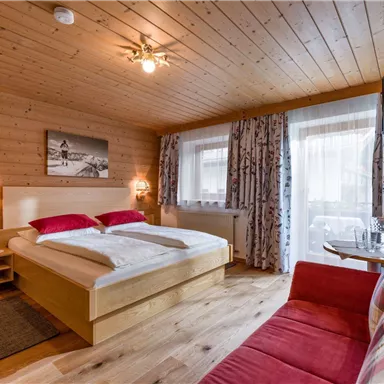 Ein gemütliches Schlafzimmer mit Holzverkleidung und großen Fenstern. Das Zimmer verfügt über ein Doppelbett, einen roten Sofa-Bereich und dekorative Vorhänge.