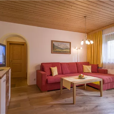 Ein gemütliches Wohnzimmer mit einem roten Sofa und einem Holztisch. Große Fenster lassen viel Tageslicht herein.