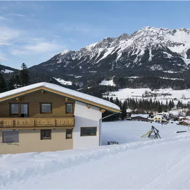 A modern house in a snowy landscape. In the background, there are tall mountains and a quiet valley town.