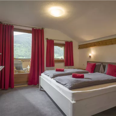 Ein gemütliches Schlafzimmer mit zwei Betten und roten Kissen. Große Fenster bieten einen Blick auf die Berge und einen Balkon.