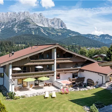 Eine wunderschöne Almhütte mit einer großen Terrasse und Liegestühlen im Garten. Im Hintergrund sind majestätische Berge und ein blauer Himmel zu sehen.