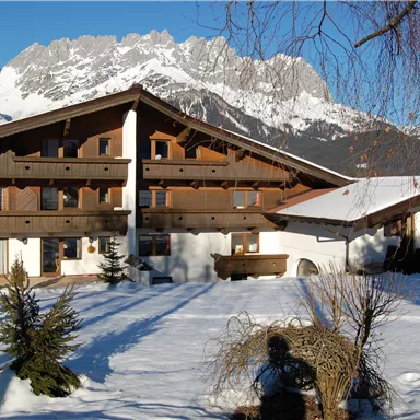 Ein skandinavisches Holzhaus im Schnee mit Bergen im Hintergrund. Die Szenerie ist ruhig und sonnig.