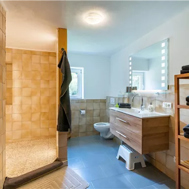 Ein modernes Badezimmer mit einer großen Dusche und einem Holzwaschtisch. Es gibt helle Fliesen und Handtücher auf einem Regal.