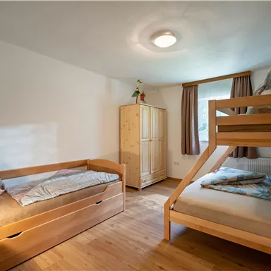Ein gemütliches Schlafzimmer mit einem Etagenbett und einem Einzelbett. Die Wände sind hell und das Zimmer hat Holzfußboden sowie eine Fensterbank mit Vorhängen.