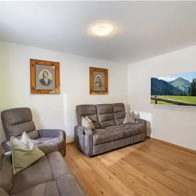 Ein gemütlicher Wohnraum mit zwei grauen Sesseln und einem modernen Sofa. An der Wand hängen nostalgische Bilder und ein Fernseher zeigt eine schöne Landschaft.