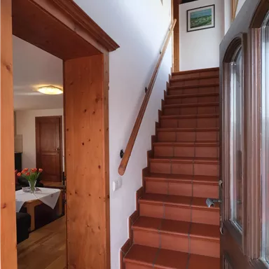 Ein Treppenhaus mit Holzstufen und hellen Wänden. Rechts befindet sich eine Eingangstür und im Hintergrund ist ein Wohnzimmer sichtbar.