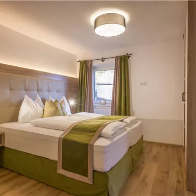 A modern bedroom with a comfortable double bed and elegant pillows. The soft lighting and green curtains create a cozy atmosphere.