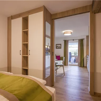 A modern bedroom with a large bed and a wardrobe. In the background, a living area with windows and a terrace is visible.