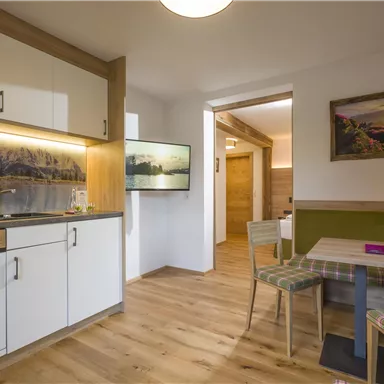 A modern kitchen with wooden fixtures and a cozy dining table. In the background, there are pictures and a television on the wall.