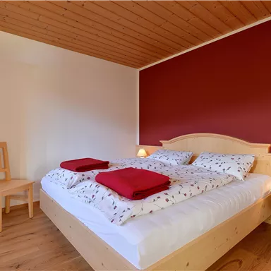 A cozy bedroom with a double bed, red pillows, and warm wood decor. The wall is painted red, and there is a chair as well as nightstands with lamps.