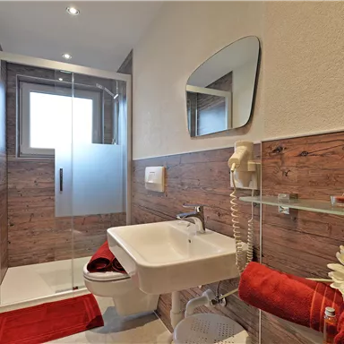 A modern bathroom with a walk-in shower and elegant wood paneling. Red towels and a flower provide a fresh accent.