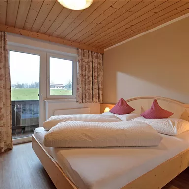 A cozy bedroom with a double bed and red pillows. The room has large windows that lead to a balcony overlooking green scenery.