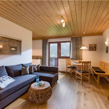 A cozy living room with a large gray sofa and wooden furniture. There is a dining area with a table and chairs as well as plenty of natural light through the window.