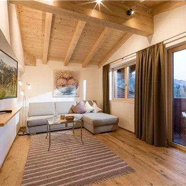 A cozy living room with wooden ceilings and warm tones. There is a sofa, a table, and access to the balcony with a view of the mountains.