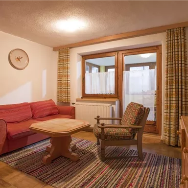 A cozy living room with a red sofa, a wooden table, and an armchair. The large windows let in plenty of daylight, and the rug creates a warm atmosphere.
