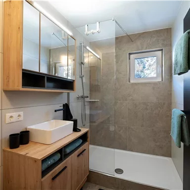 A modern bathroom with a glass shower and wooden shelves. The decor is simple and elegant, featuring a large mirror and a sink on a wooden cabinet.