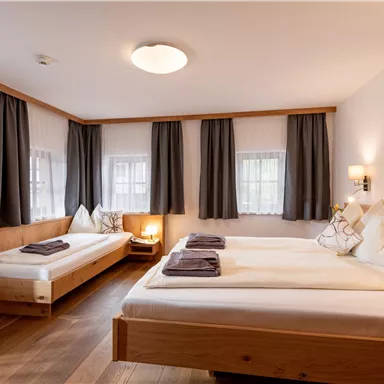 A cozy room with two beds, an additional bed, and wooden details. The windows are draped with curtains and there is warm light from a lamp.