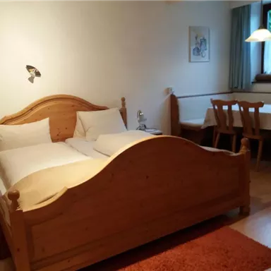 A cozy bedroom with a wooden bed and two pillows. Next to the bed is a dining table with chairs and a window that lets in daylight.