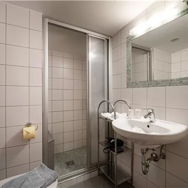 A modern bathroom with white tiles, a shower, and a sink. There is a mirror and a towel holder.