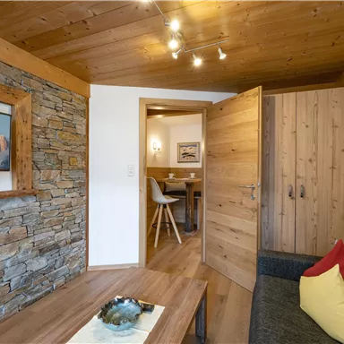 A cozy room with a wooden ceiling and stone walls. In the background, an adjacent dining area is visible.