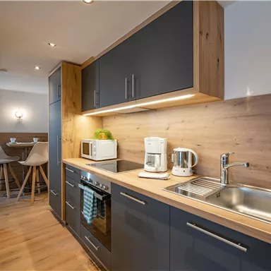 A modern kitchen with dark cabinets and wooden details. It includes appliances such as a microwave, coffee machine, and a stove.