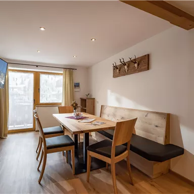 A bright dining room with a wooden table and chairs. A television hangs on the wall, and the windows let in plenty of light.