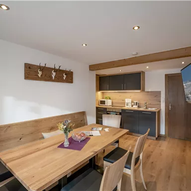 A cozy room with a wooden dining table and modern furniture. In the corner is a small kitchenette and a television on the wall.