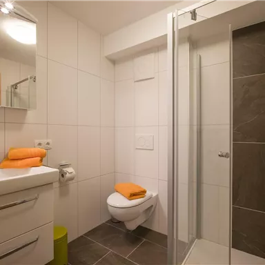 Ein modernes Badezimmer mit einer Dusche und einem WC. Auf dem Waschbecken liegen orangefarbene Handtücher.