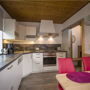 Eine moderne Küche mit weißen Möbeln und Holzdetails. Der Raum ist gut beleuchtet und hat einen Esstisch mit roten Stühlen.